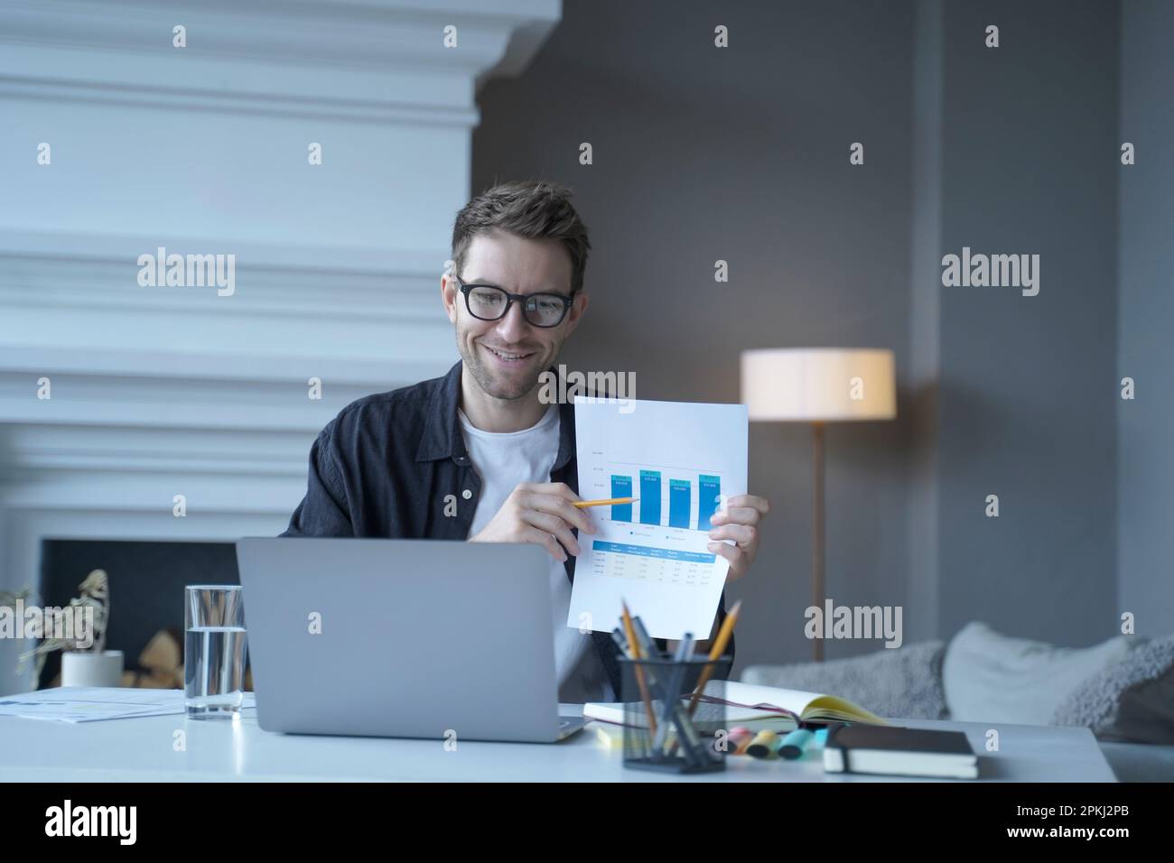 Giovane uomo d'affari tedesco sorridente in abiti casual che punta a documento con grafica durante la riunione su Internet e parlare con i colleghi on-line, mentre Foto Stock