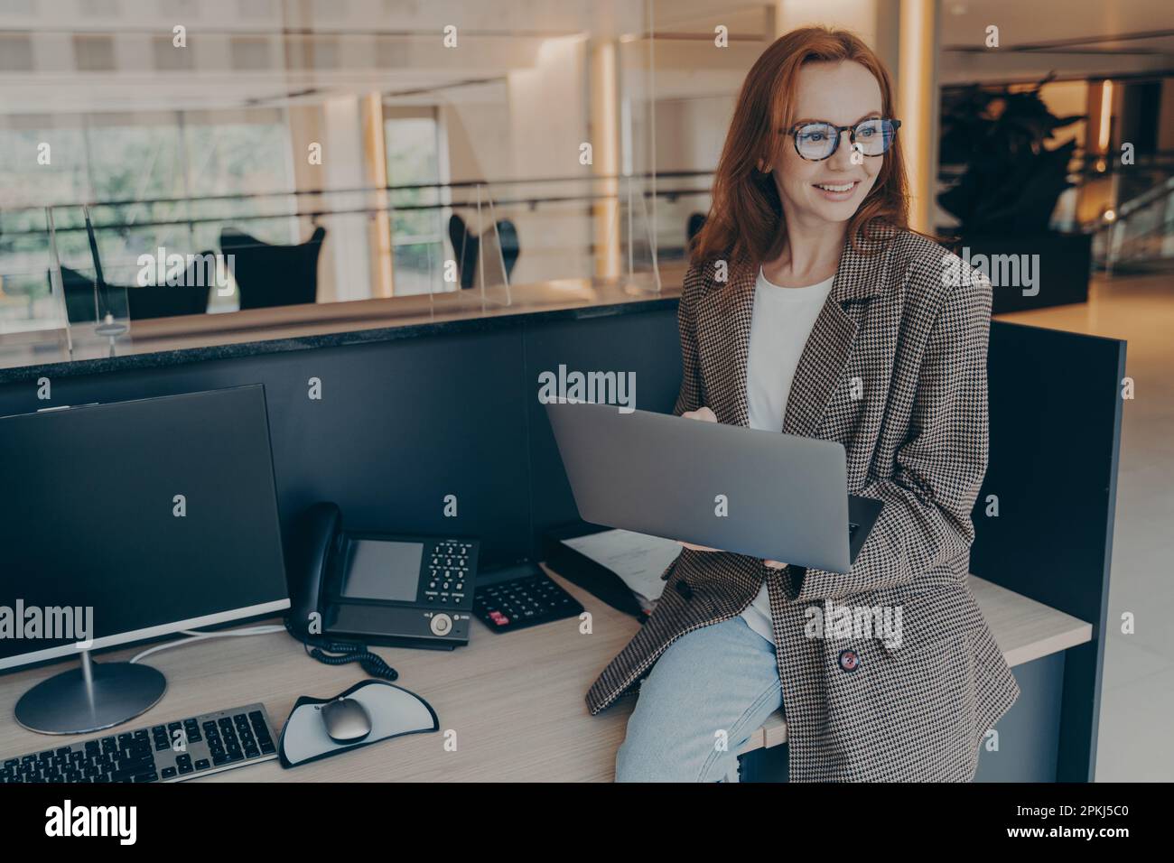 Sorridente dipendente di un ufficio con capelli rossi in occhiali seduti sulla scrivania con varie attrezzature mentre tiene in mano il computer portatile, si guarda intorno e si naviga sul Web con la collaborazione Foto Stock