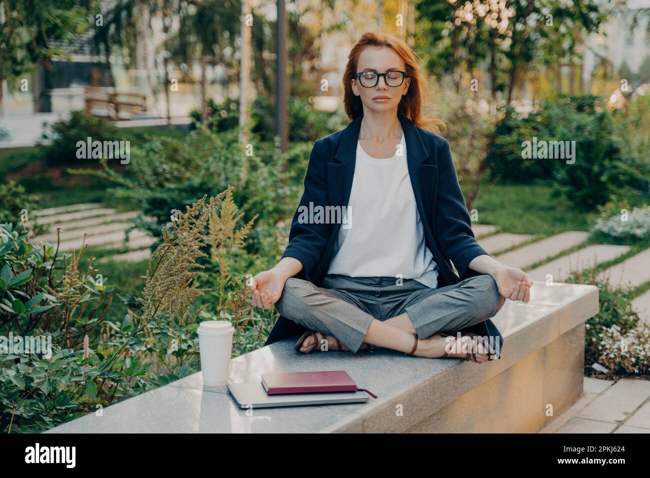 Realxed redhead donna fa yoga fuori al parco si siede in lotus posa medita all'aperto indossa occhiali formale vestito raccoglie con pensieri prima del lavoro Foto Stock