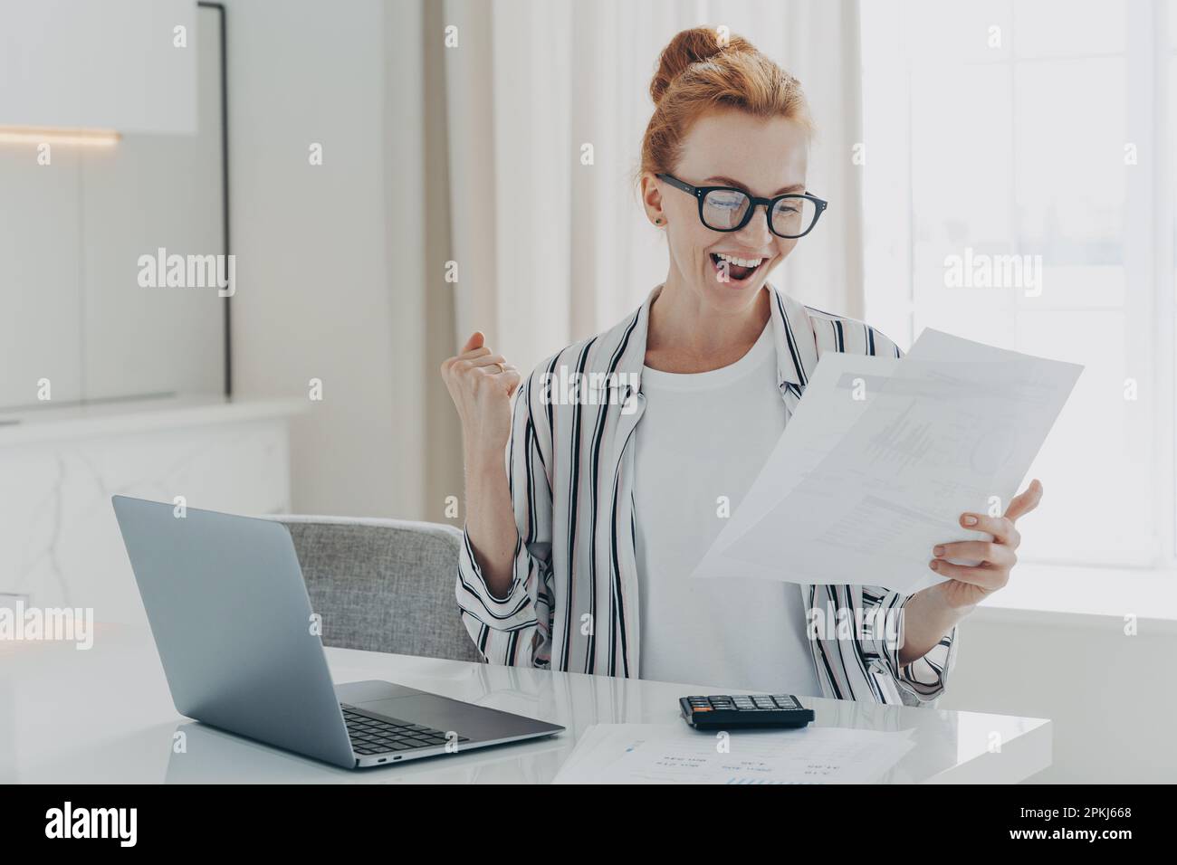 Emotivo felice giovane donna zenzero in eyewear tenendo carta con l'ultima notifica bancaria pagamento ipotecario, sollevando la mano con pugno clenched, lettura Foto Stock