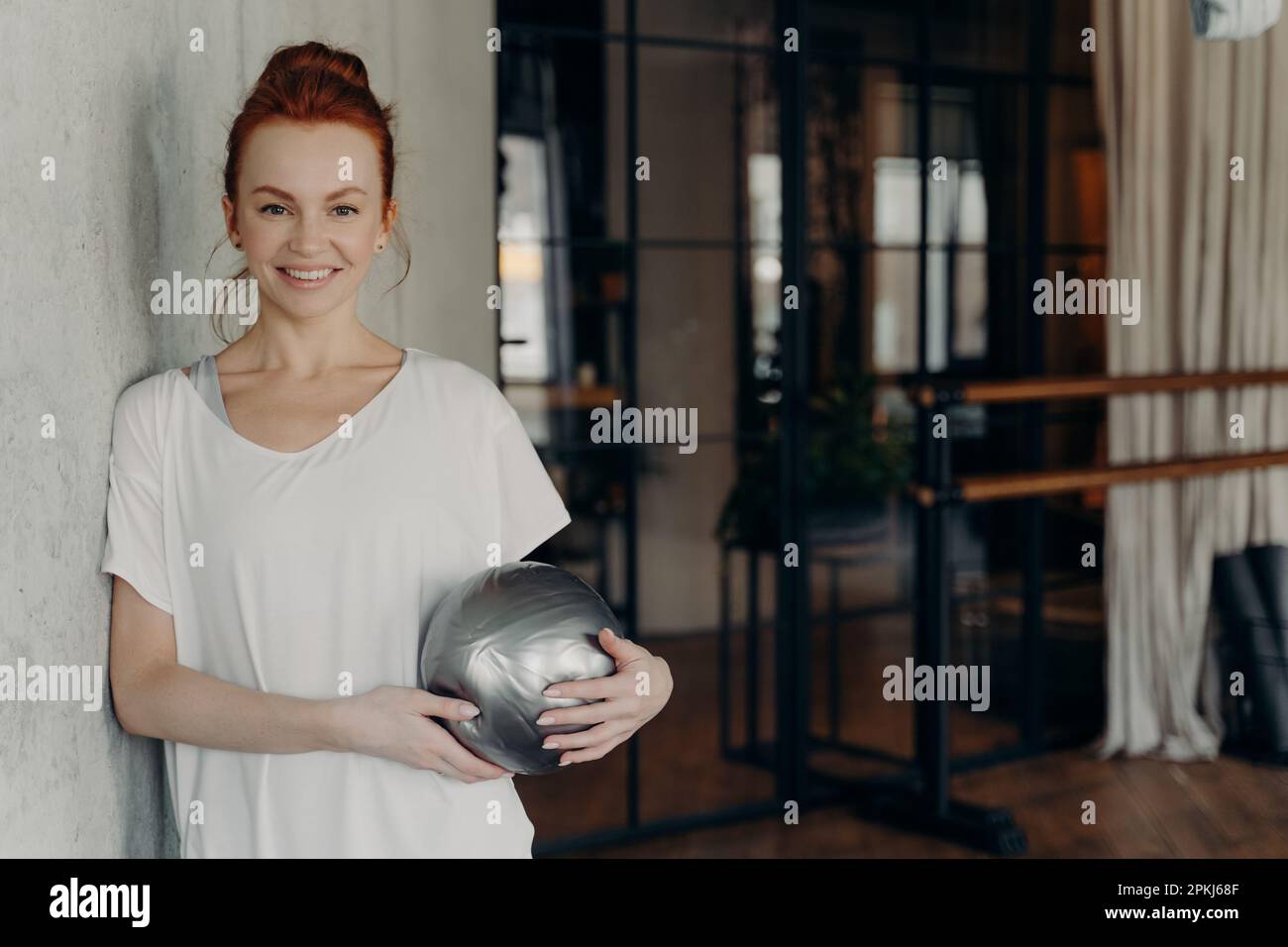 Giovane bella donna in forma con sorriso con palla mini dimensione guardando positivamente la macchina fotografica mentre si appoggia su parete di cemento grigio nel centro fitness ambienc Foto Stock