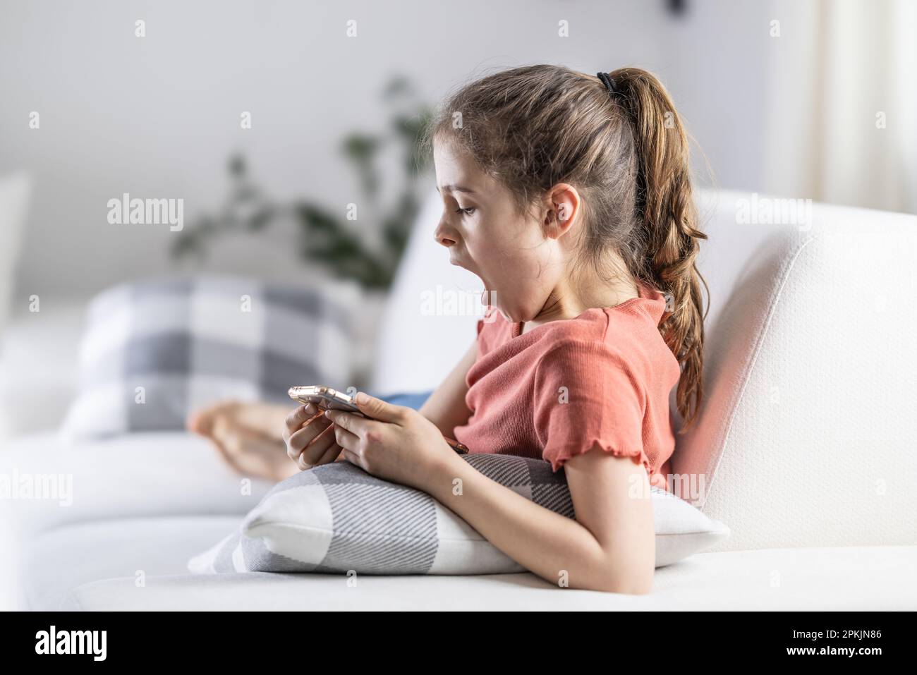 La ragazza che guarda nel suo telefono si sente stanca o annoiata mentre si siede a casa su un divano. Foto Stock