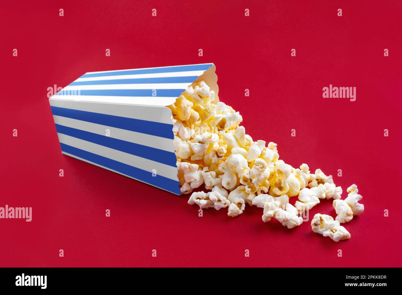 Gustoso popcorn al formaggio che cade da un secchio di cartone a strisce blu, isolato su sfondo rosso. Dispersione di grani di popcorn. Film, cinema e intrattenimento Foto Stock