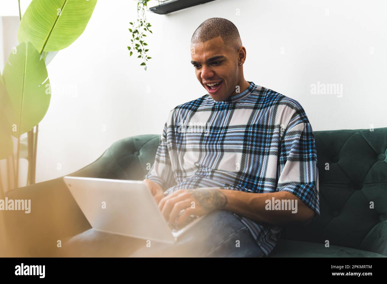 Il giovane afro-americano tatuò un uomo che guardava qualcosa che non si aspettava su un computer portatile mentre era seduto su un divano a casa. Foto di alta qualità Foto Stock