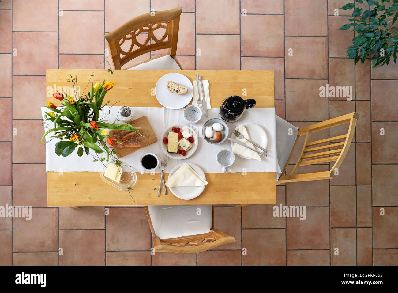 Colazione su un tavolo di legno con pane, formaggio, uova e caffè e un bouquet di tulipani come decorazione, tre sedie d'epoca su un pavimento piastrellato, alto ang Foto Stock