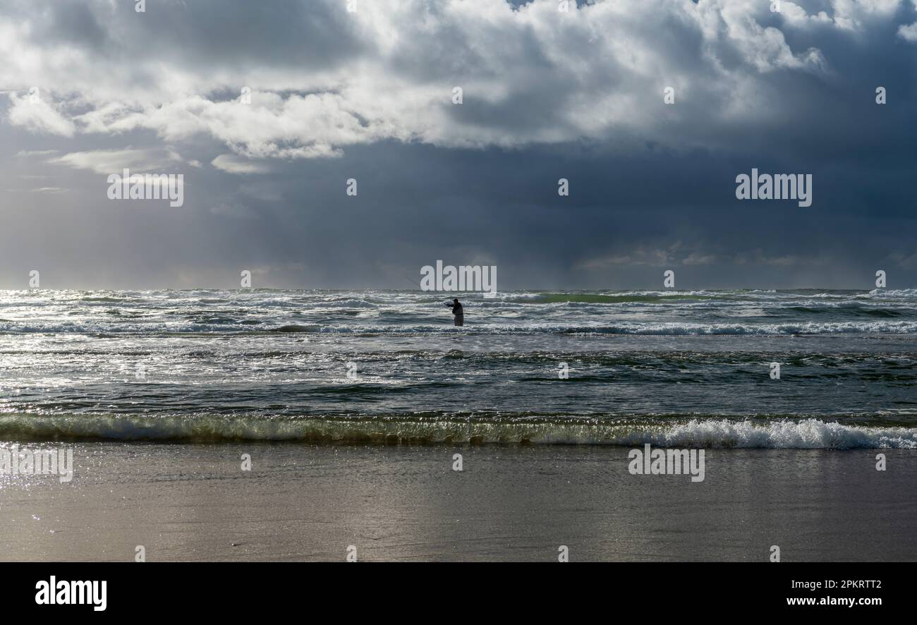 Un pescatore da solo che indossa una muta in piedi in acque oceaniche con bassa marea e retroilluminata dalla luce del tramonto Foto Stock