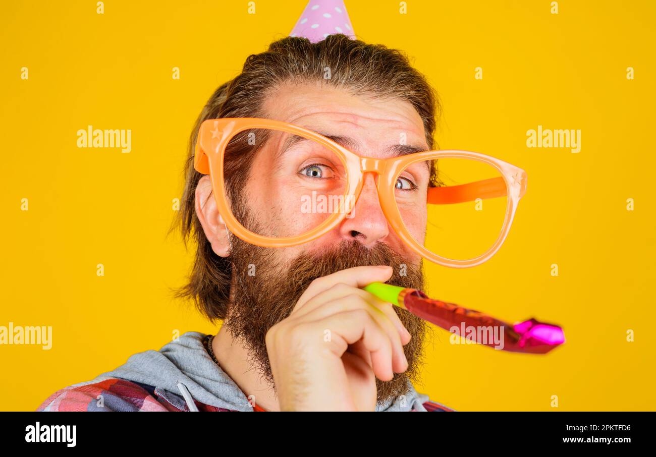 Buon compleanno. Uomo bearded in cappello di compleanno e grandi bicchieri che soffia fischio partito. Bell'uomo in cappello conico e divertenti occhiali che fischiano nel corno del partito Foto Stock