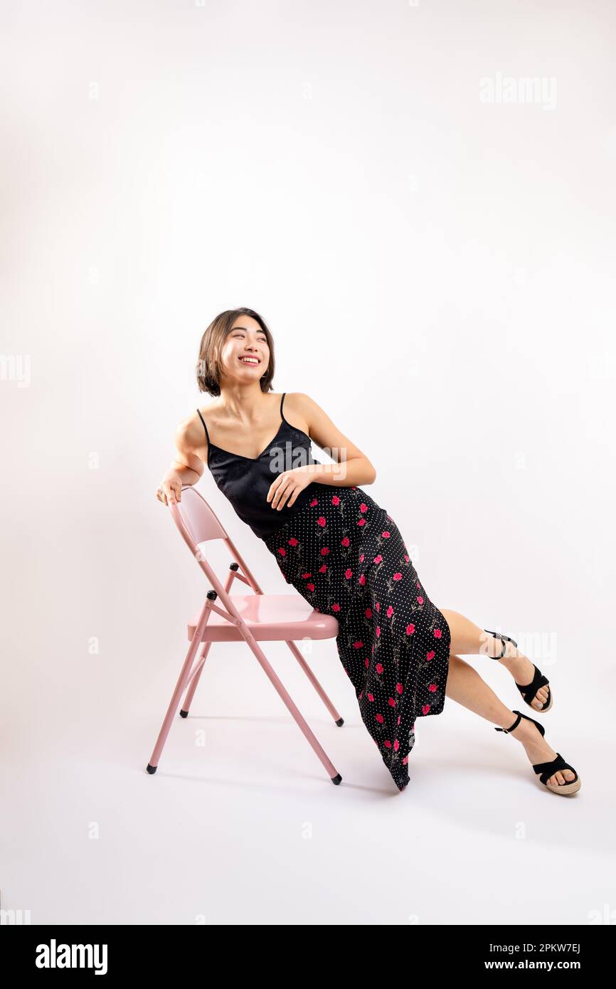Giovane donna asiatica seduta su una sedia rosa in Studio | sfondo bianco | Abito con stampa floreale | Top in seta nera Foto Stock