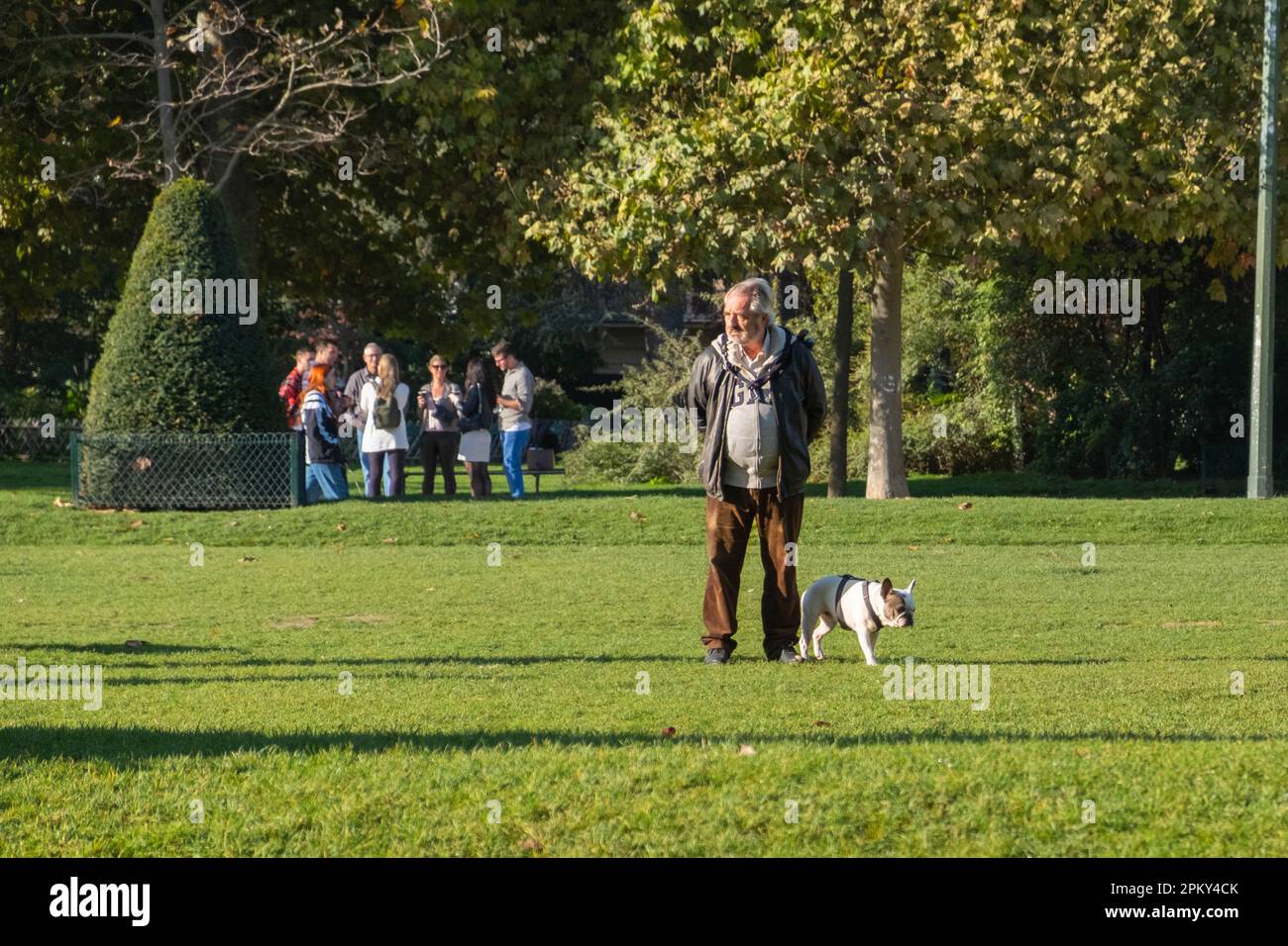 Un uomo di mezza età che cammina con il suo bulldog francese in una giornata di sole in un lussureggiante giardino di Parigi, godendosi la pace all'aria aperta e il legame con il suo animale domestico. Foto Stock