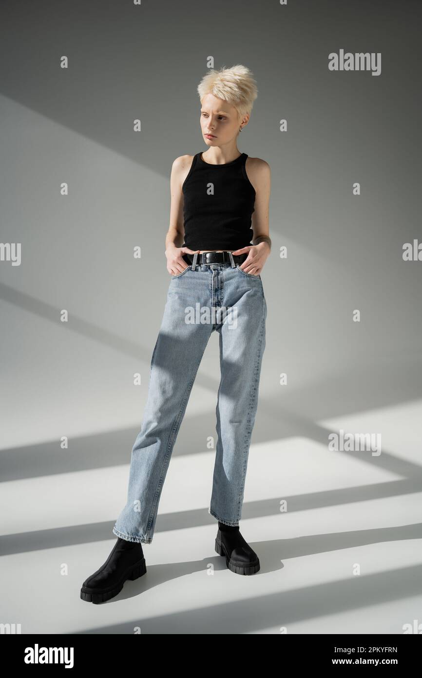 intera lunghezza di donna albino in nero canotta top e jeans posa con le mani in tasche su sfondo grigio Foto Stock
