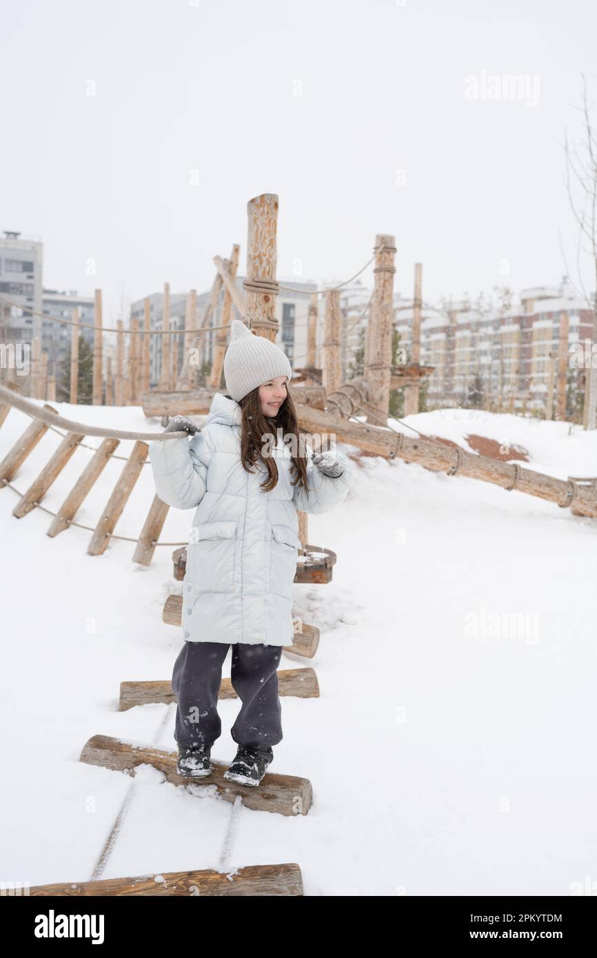 Vista laterale del corpo della giovane ragazza in abiti caldi, in piedi sulla scala di tronchi nel parco giochi innevato Foto Stock