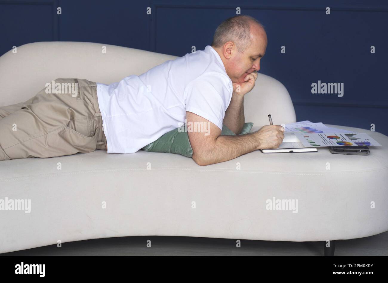 Un uomo d'affari si trova su un divano e guarda le tendenze dei dati su grafici e grafici, prende appunti in un notebook. Lavoro a distanza Foto Stock