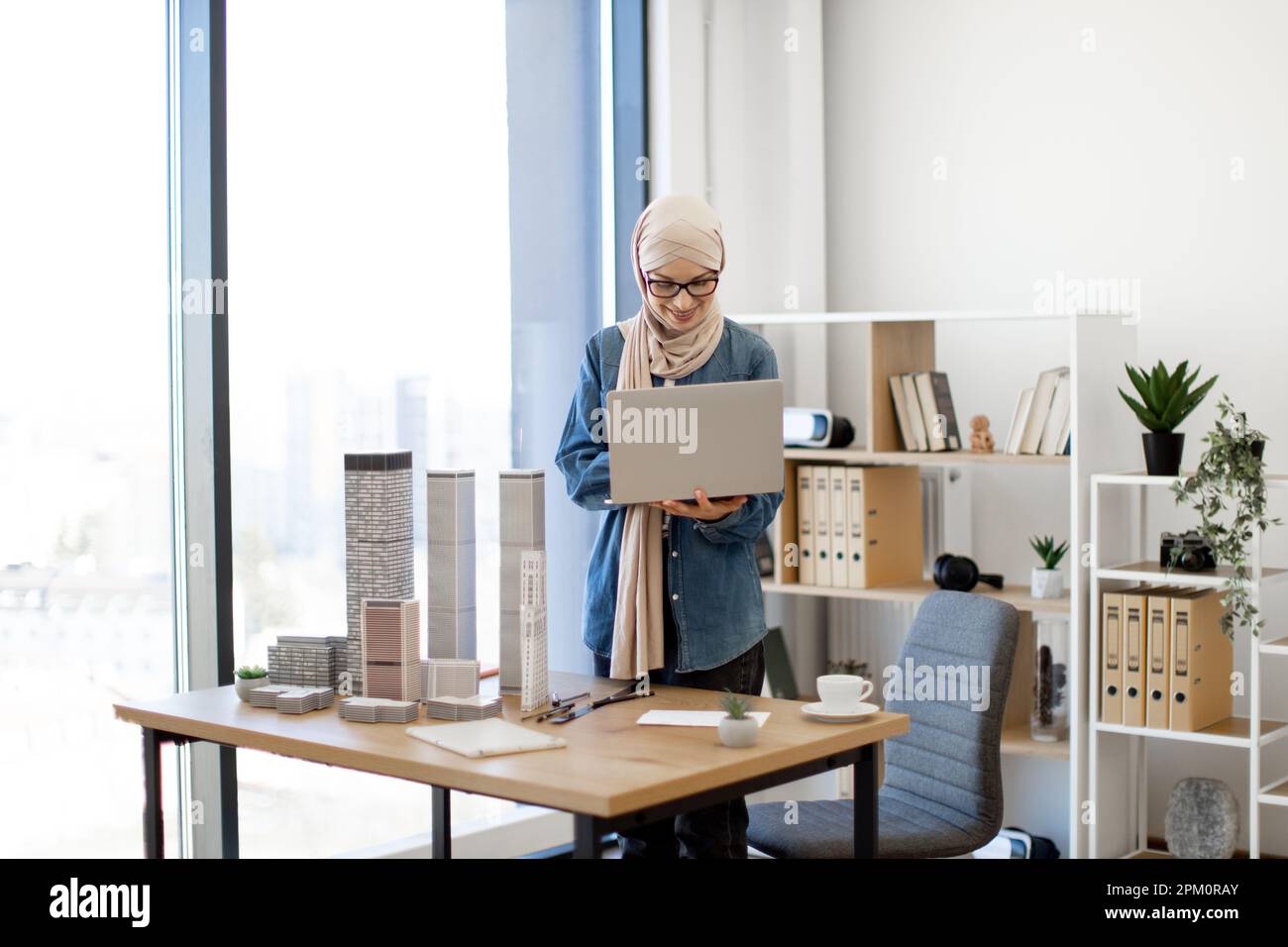 Allegra donna musulmana in hijab che gestisce un moderno computer portatile mentre si trova dietro la scrivania con edificio complesso prototipo in ufficio. Design professionale creazione di progetti architettonici al lavoro. Foto Stock