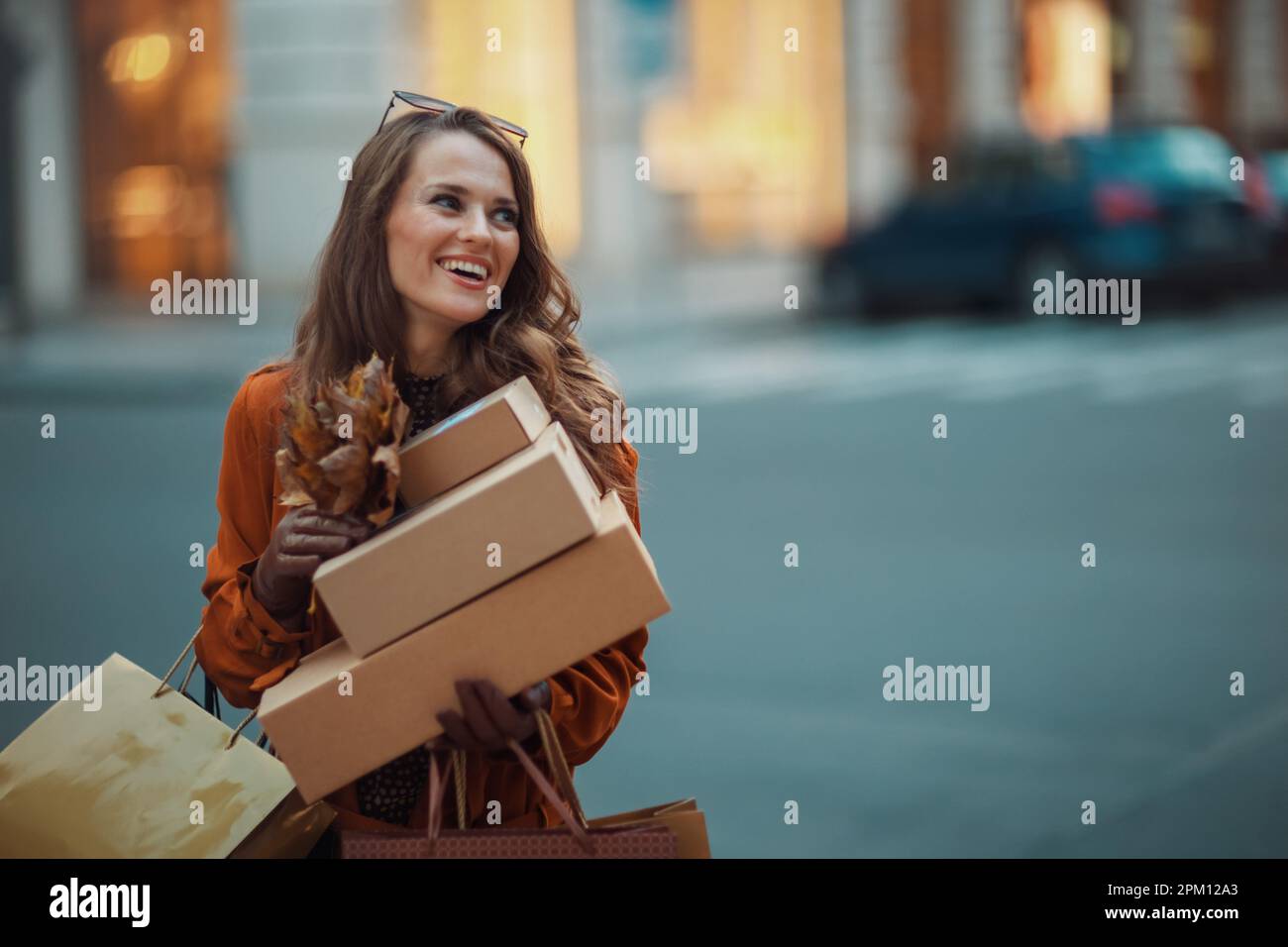 Ciao settembre. donna sorridente moderna di 40 anni in trench marrone con pacchi, borse per la spesa e foglie gialle autunnali in città. Foto Stock