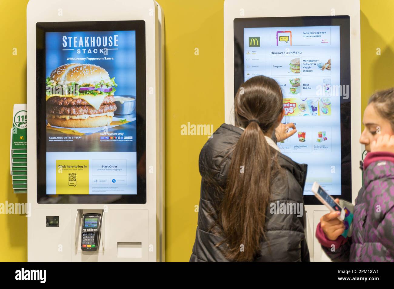 Le donne clienti che ordinano cibo su un sistema di ordinazione del touch screen LED al ristorante McDonald's, un altro schermo che mostra STEAKHOUSE Stack England UK Foto Stock
