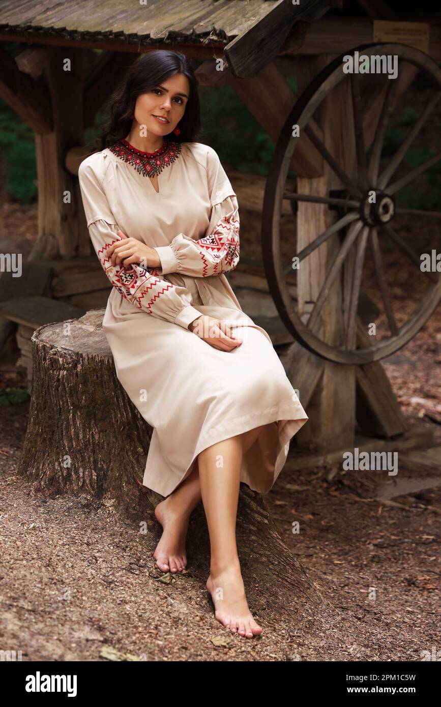 Bella donna con vestito ricamato seduta vicino al vecchio pozzo di legno in campagna. Vestiti nazionali ucraini Foto Stock