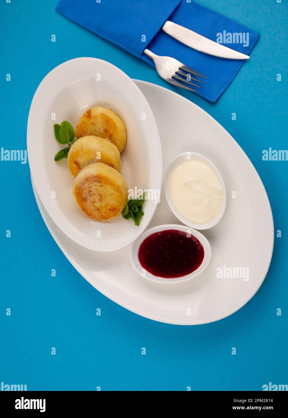 Frittelle di formaggio ricotta rotonda in un piatto bianco su sfondo blu. Cibo prima colazione. Cibo sano. Salsa di panna acida e menta. Primo piano vista dall'alto. Pasto dessert. Frittelle di ricotta al formaggio ricotta. Foto Stock
