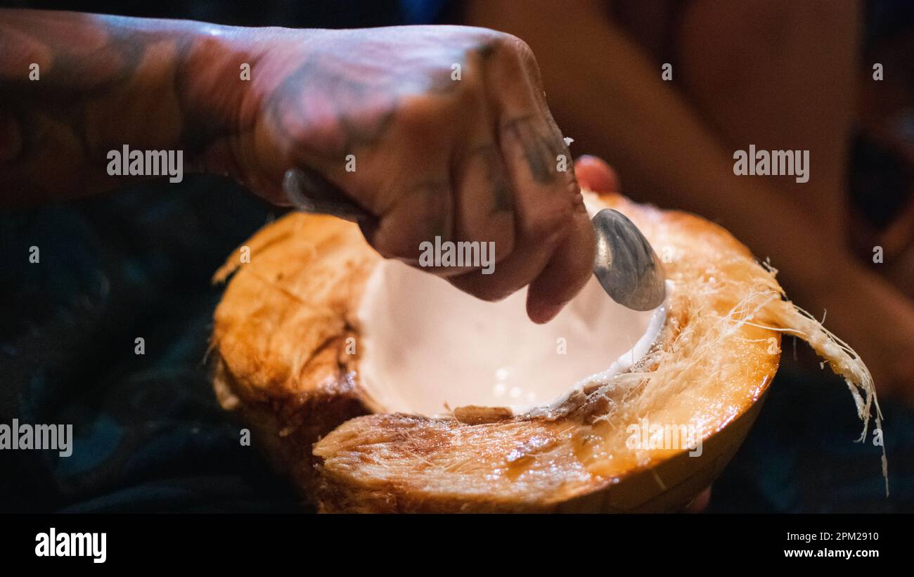 Mani tatuate che raccolgono carne fresca di cocco da una noce di cocco dimezzata in uno scatto di dettaglio ravvicinato, Filippine, Sud-est asiatico. Foto Stock