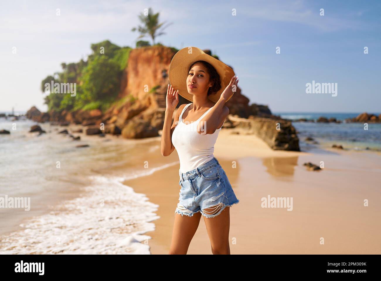 Giovane donna africana modello in cappello di paglia e occhiali da sole in posa presso il resort dal mare all'alba. Donna nera contro l'isola rocciosa e l'oceano Foto Stock