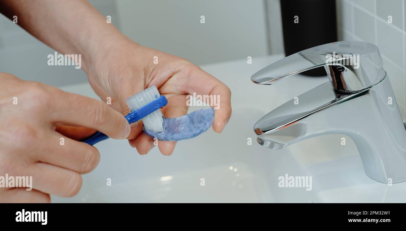 un uomo pulisce la sua stecca occlusale blu usando uno spazzolino da denti blu in un lavandino del bagno, in un formato panoramico da usare come banner web o intestazione Foto Stock