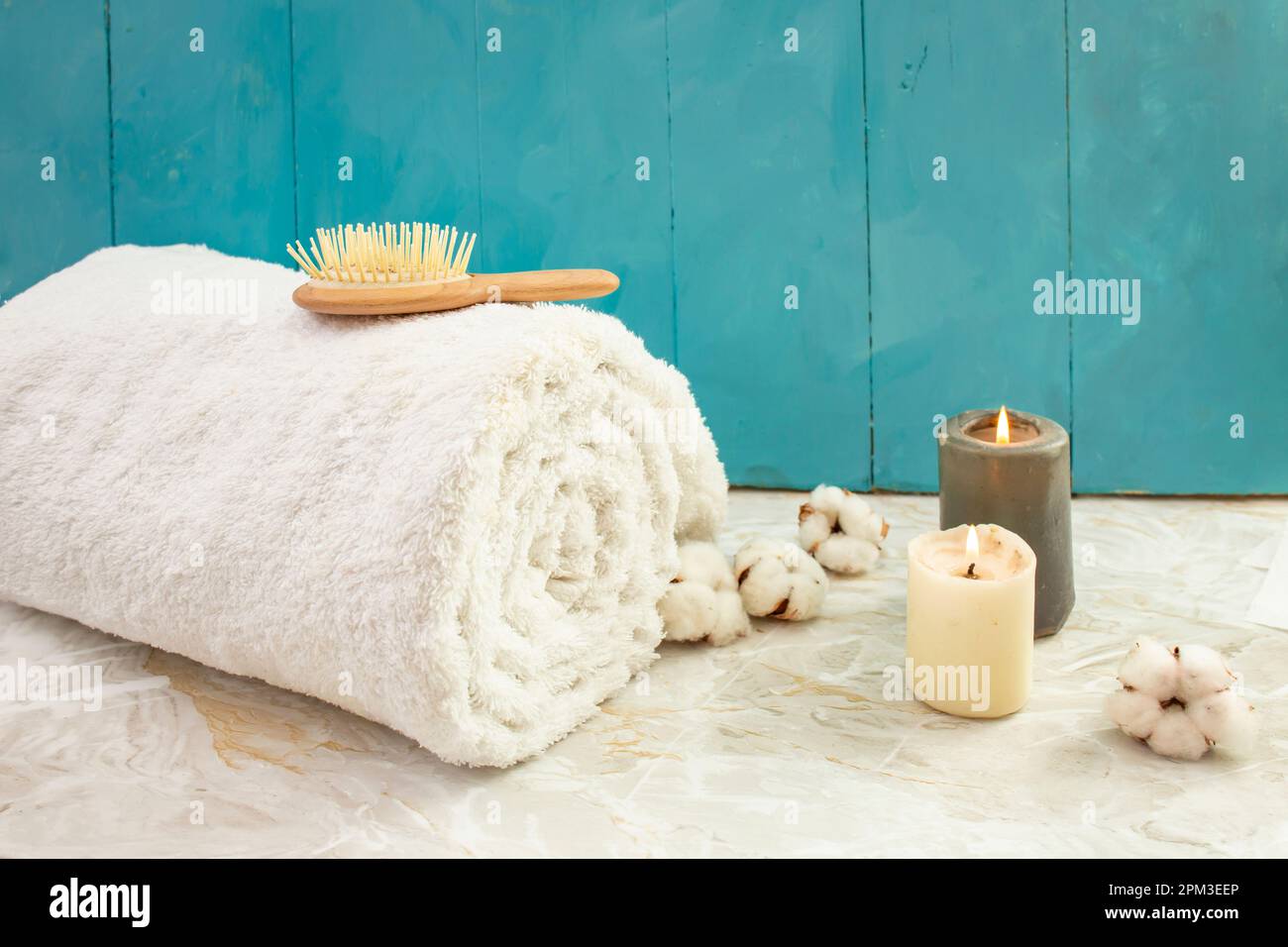 Asciugamano bianco arrotolato, spazzola per capelli di legno, fiori di cotone e candele accese su tavole di legno blu e sfondo di marmo Foto Stock