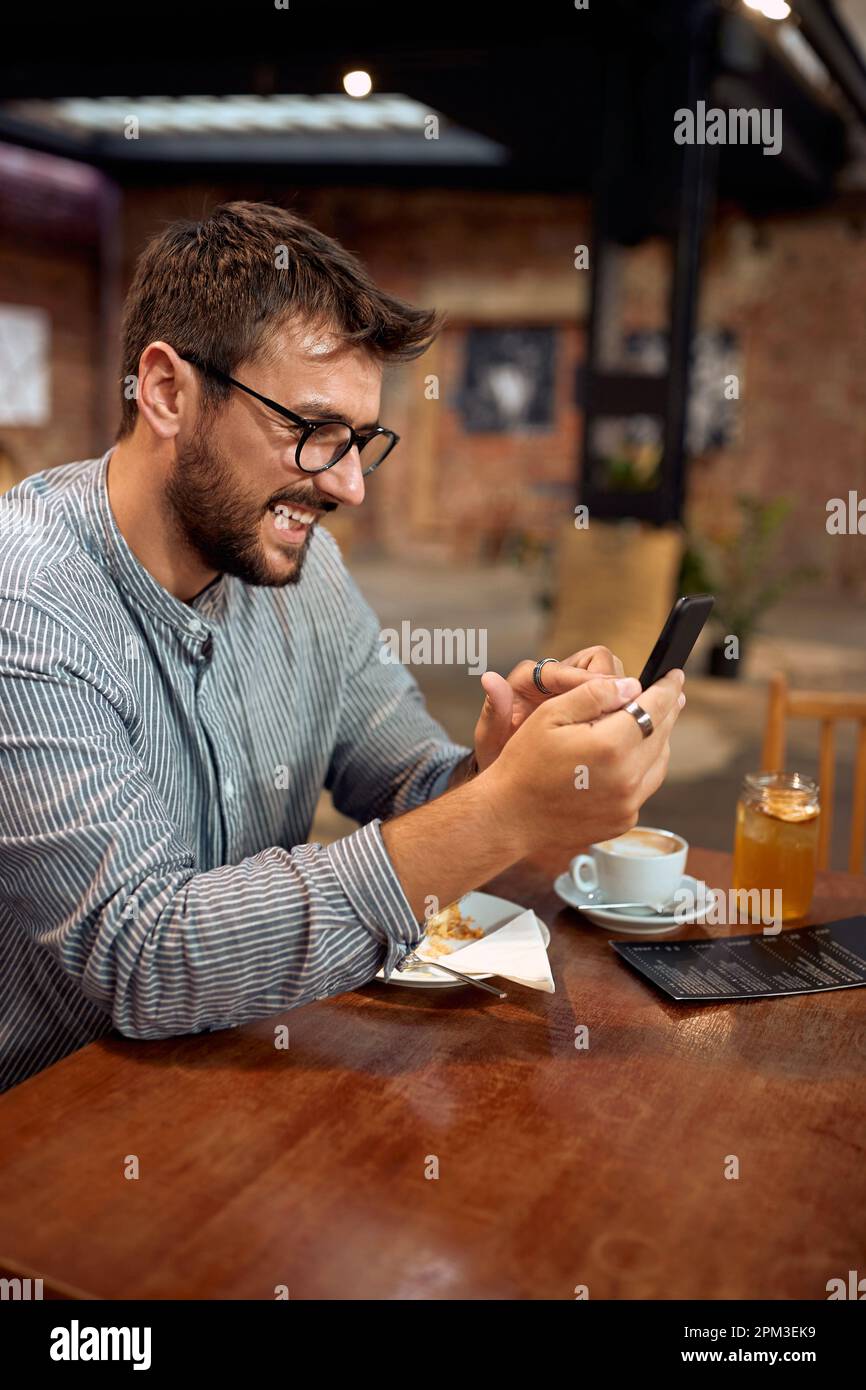 Bel giovane uomo al bar, seduto da solo al tavolo che scorre sul suo telefono, godendo caffè, torta e succo di frutta. Concetto di studente, business, lifestyle. Foto Stock