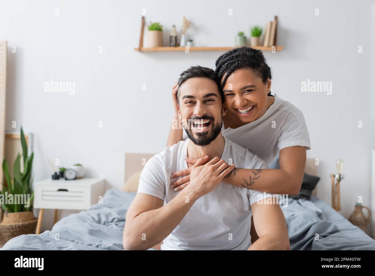 coppia multietnica overjoyed in t-shirt bianche ridendo alla macchina fotografica in camera da letto a casa, immagine stock Foto Stock
