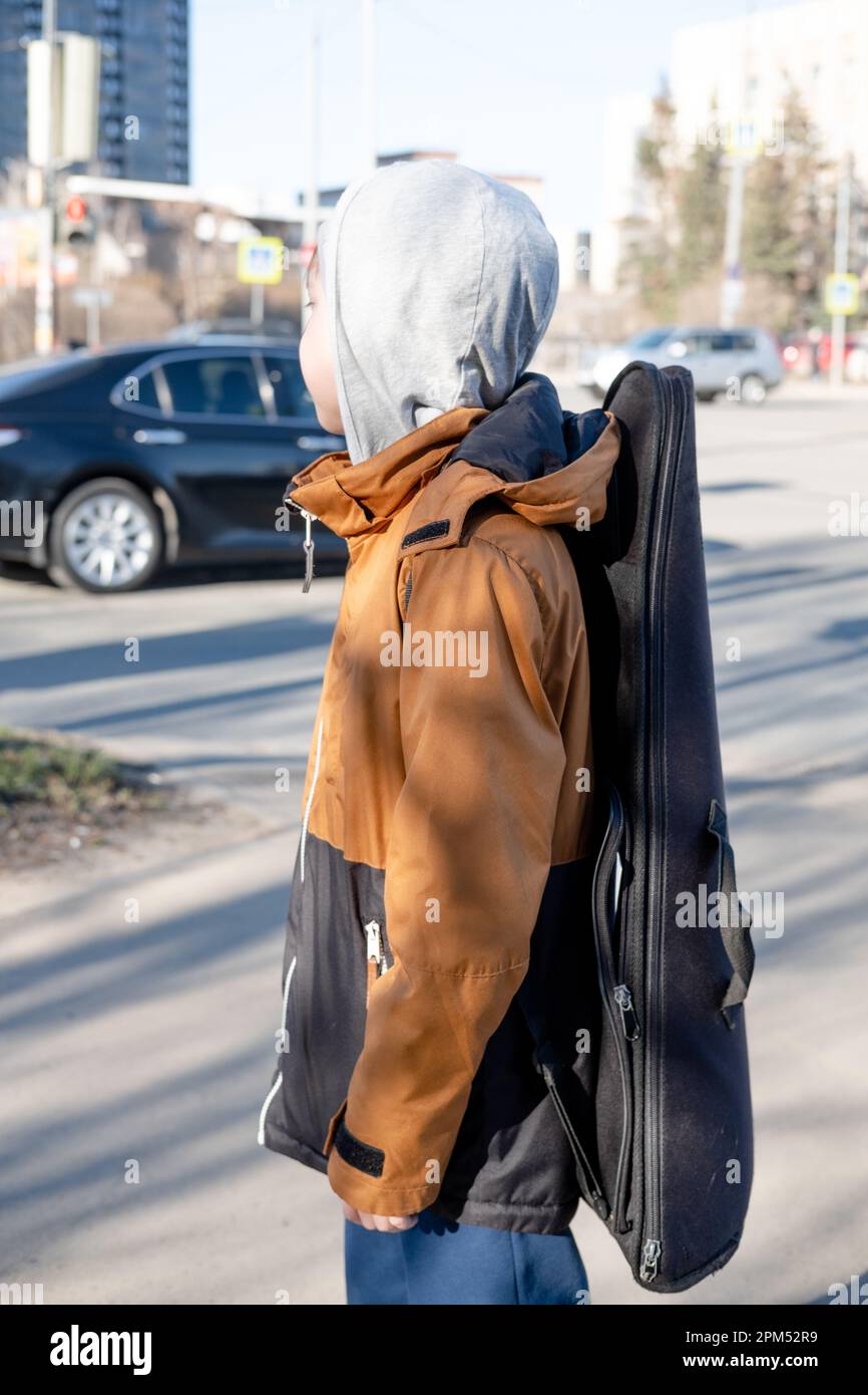 Un ragazzo adolescente in giacca e cappello cammina da solo con una chitarra in un astuccio. Musicista bambino perso in città Foto Stock