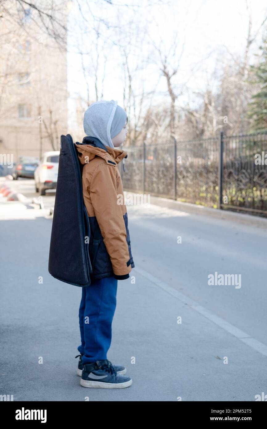 Un ragazzo adolescente in giacca e cappello cammina da solo con una chitarra in un astuccio. Musicista bambino perso in città Foto Stock