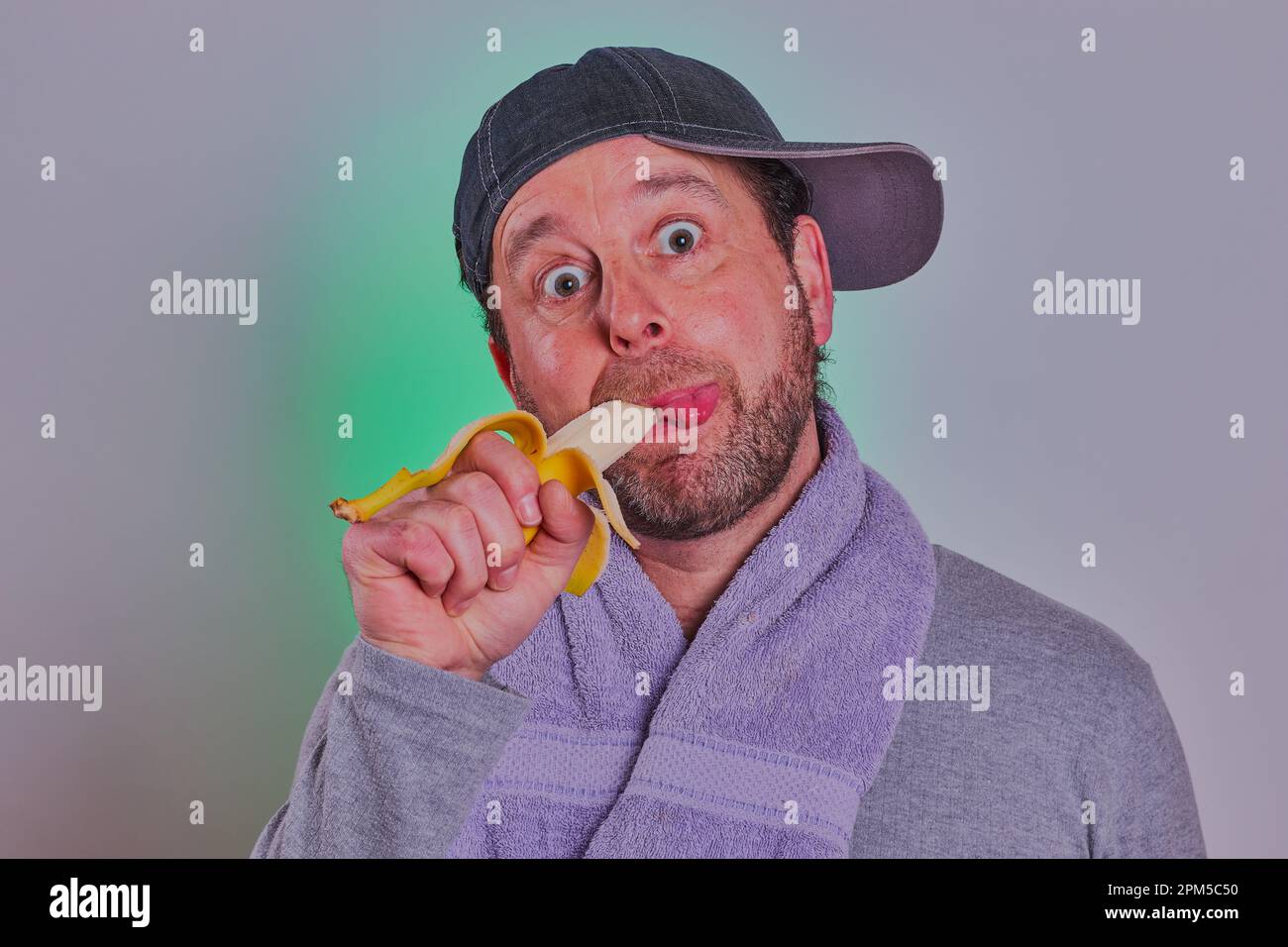 Uomo barbuto con base a sfera e asciugamano intorno al collo che mangia una banana. Modello disponibile. Foto Stock