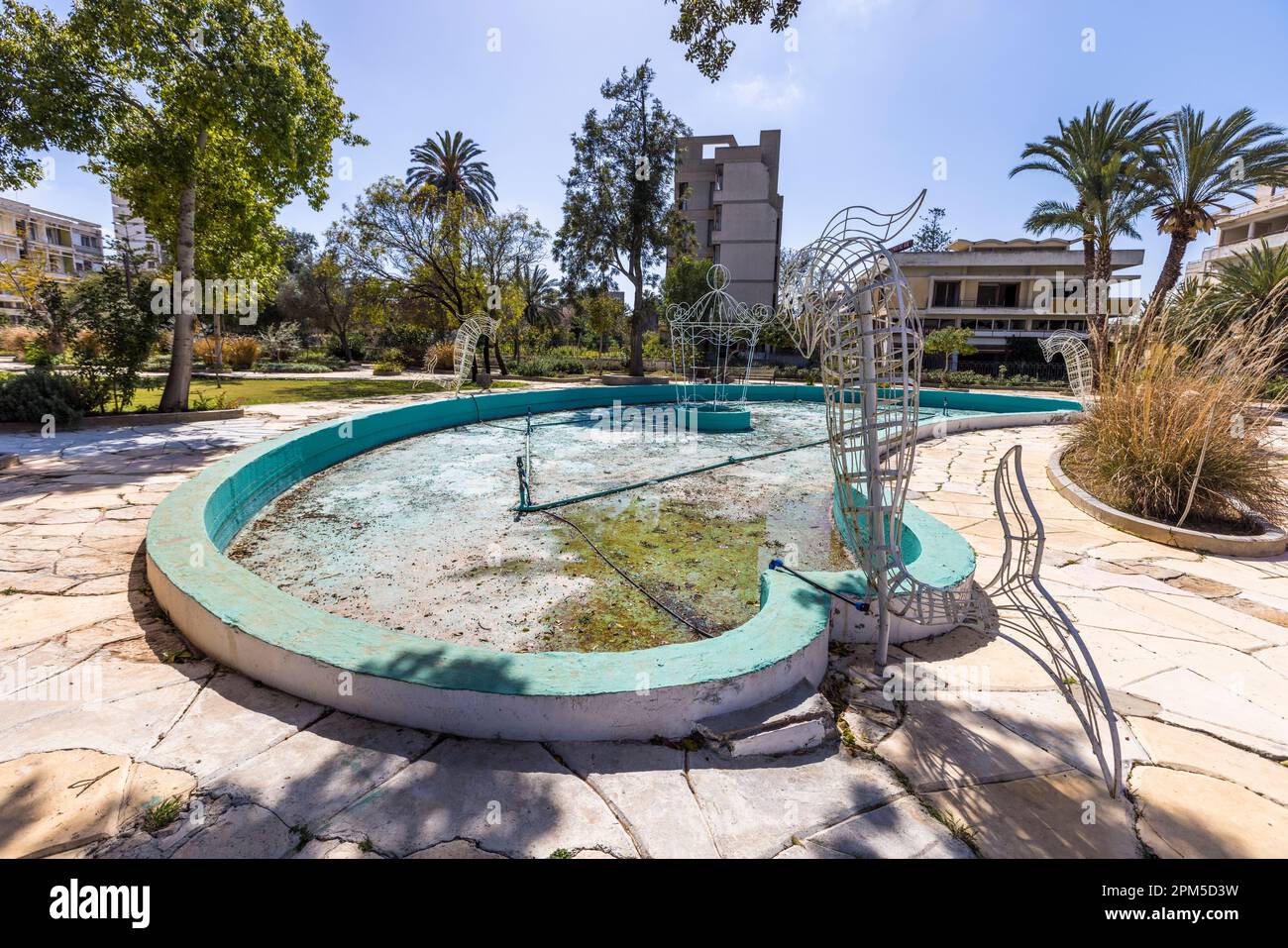 Un bacino di fontana secco e macchiato di alghe con sculture di pesci arrugginite giace in un parco invaso dalla vegetazione nella città fantasma di Varosha vicino a Famagusta, Repubblica Turca di Cipro del Nord (RTNC). Una torre d'albergo vuota si erge tra le palme sullo sfondo. Fino all'invasione turca del 1974, Varosha era una località balneare glamour con il jet set internazionale; da allora è una zona militare riservata e una piccola parte è aperta ai visitatori dalla fine del 2020. Foto Stock
