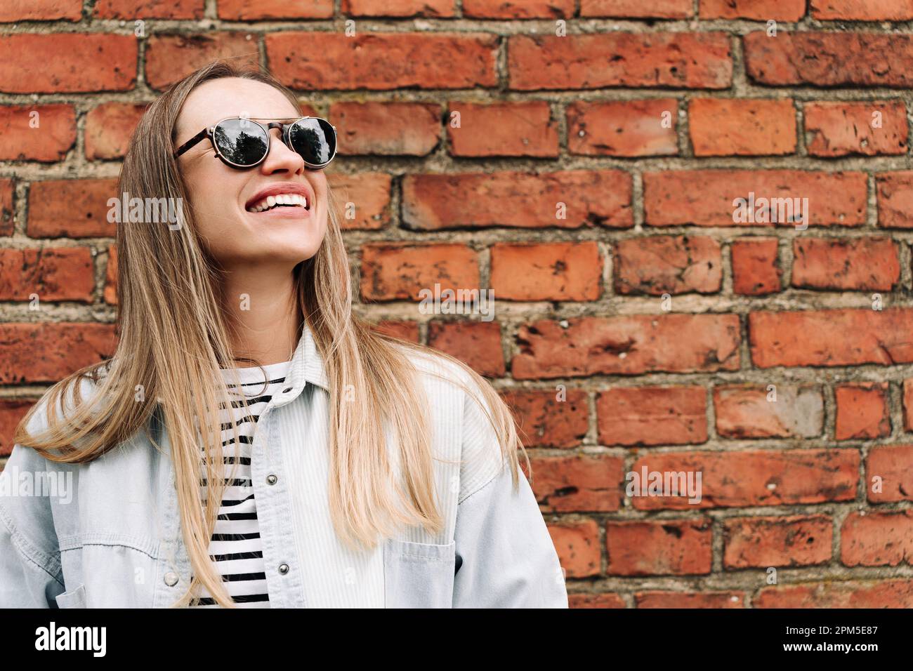 Donna dai capelli lunghi in occhiali da sole ride vicino al muro di mattoni Foto Stock