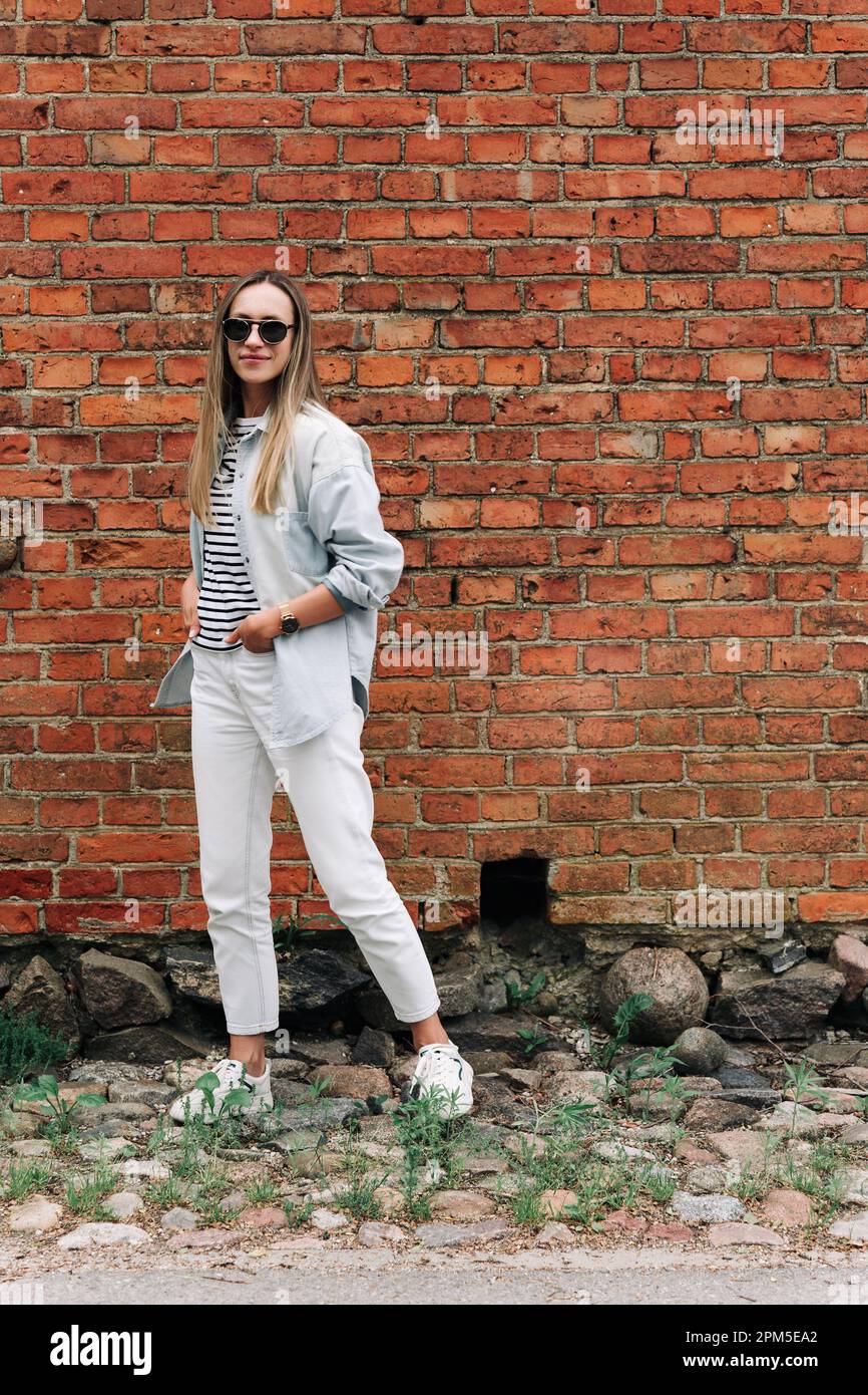 Una donna in abiti leggeri e occhiali da sole si trova vicino a una parete di mattoni Foto Stock
