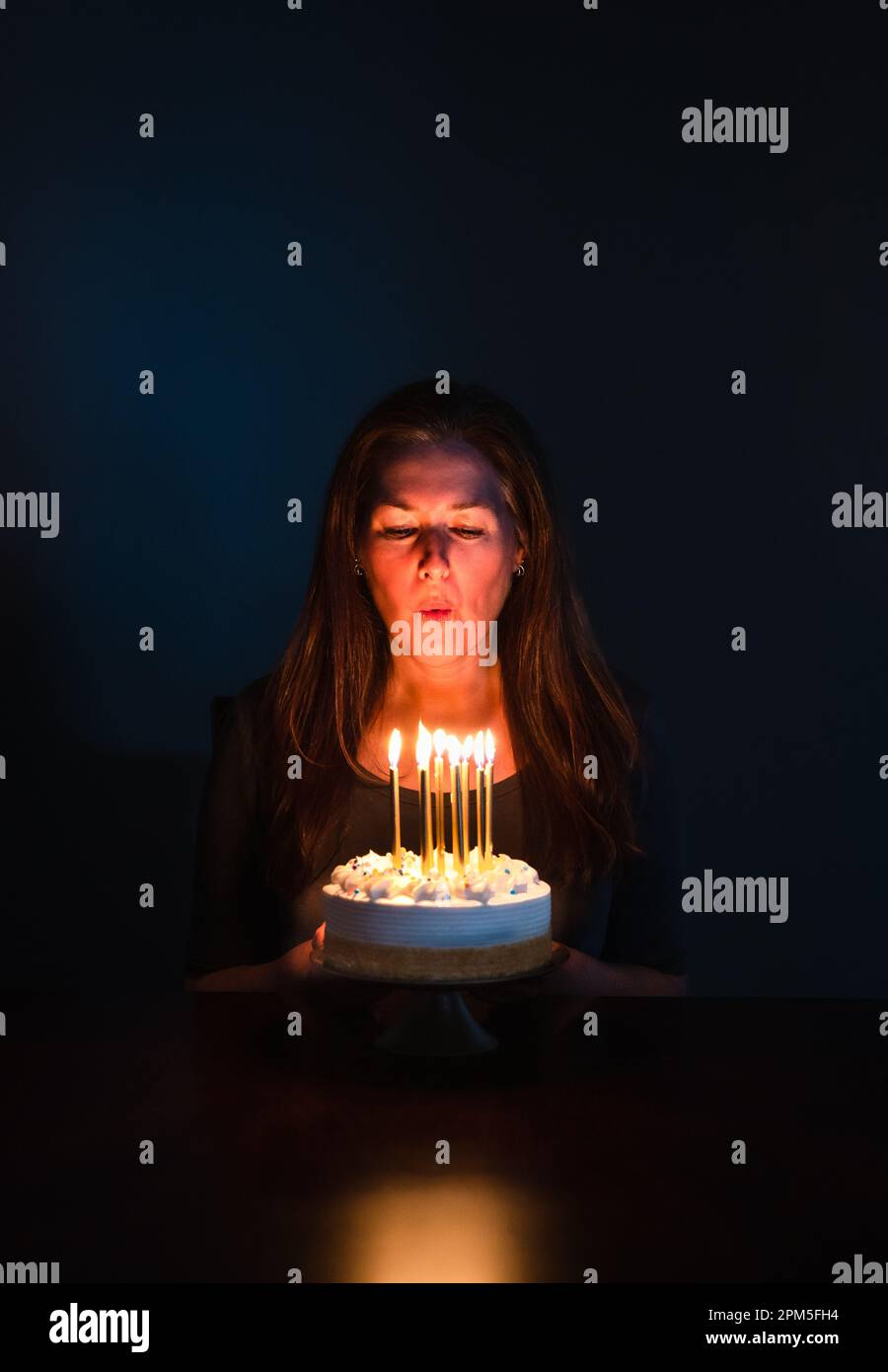 Donna attraente che soffia le candele sulla torta di compleanno in camera scura. Foto Stock