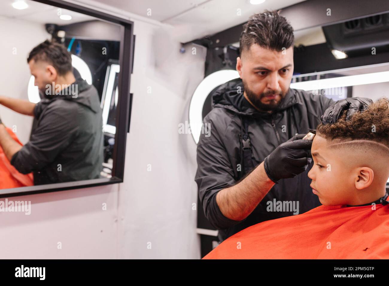 Il barbiere latino si è concentrato allineando i bordi misti dei ragazzi durante il taglio dei capelli Foto Stock