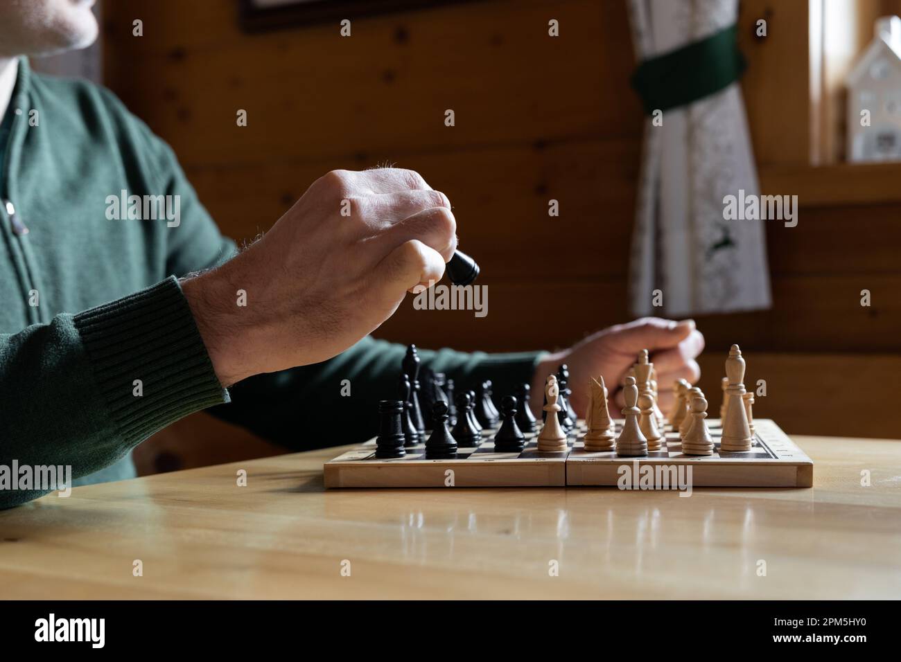 Un uomo gioca a scacchi su un tavolo di legno vicino ad una finestra e tiene un pezzo nero nelle sue mani. Foto Stock