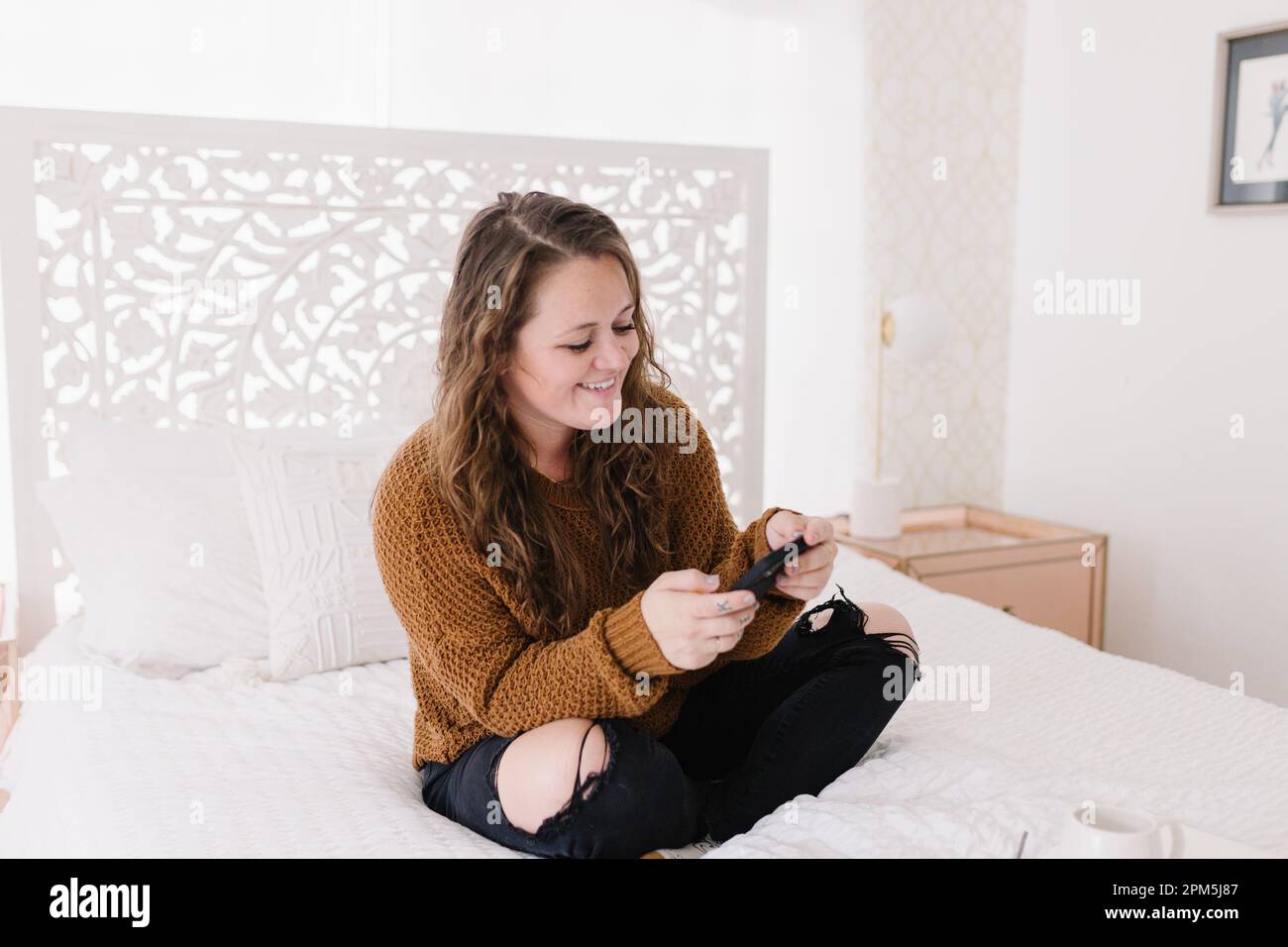 Felice giovane donna guardando il telefono seduto sul letto bianco Foto Stock