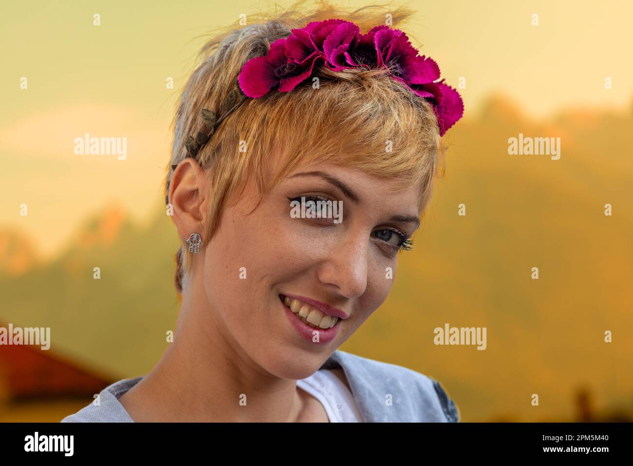 Primo piano ritratto di una donna con capelli biondi corti, ordinato e fatto, indossando un archetto fatto di fiori di fucsia, su uno sfondo esterno che Foto Stock