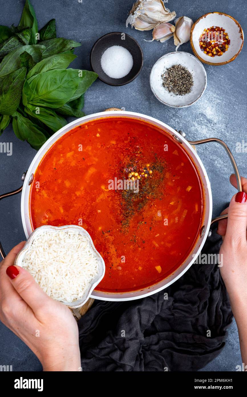mani di donna aggiungendo riso in zuppa di pomodoro che sta cucinando in una pentola, spezie e foglie di basilico sul lato. Foto Stock