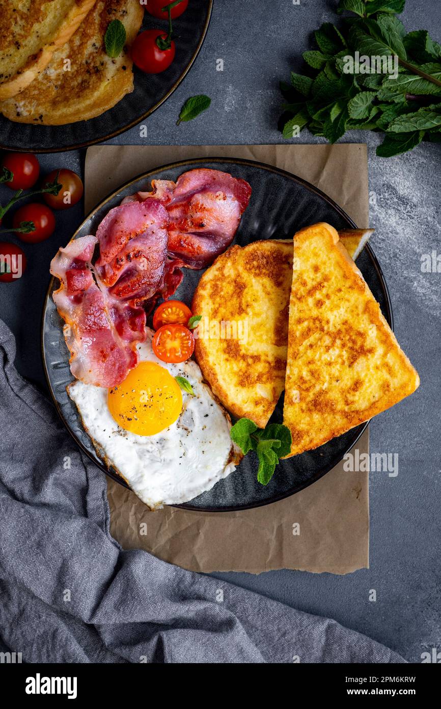 Un piatto per la colazione con toast alla francese, uova fritte e pancetta guarnita con foglie di menta fresca e pomodori ciliegini. Foto Stock