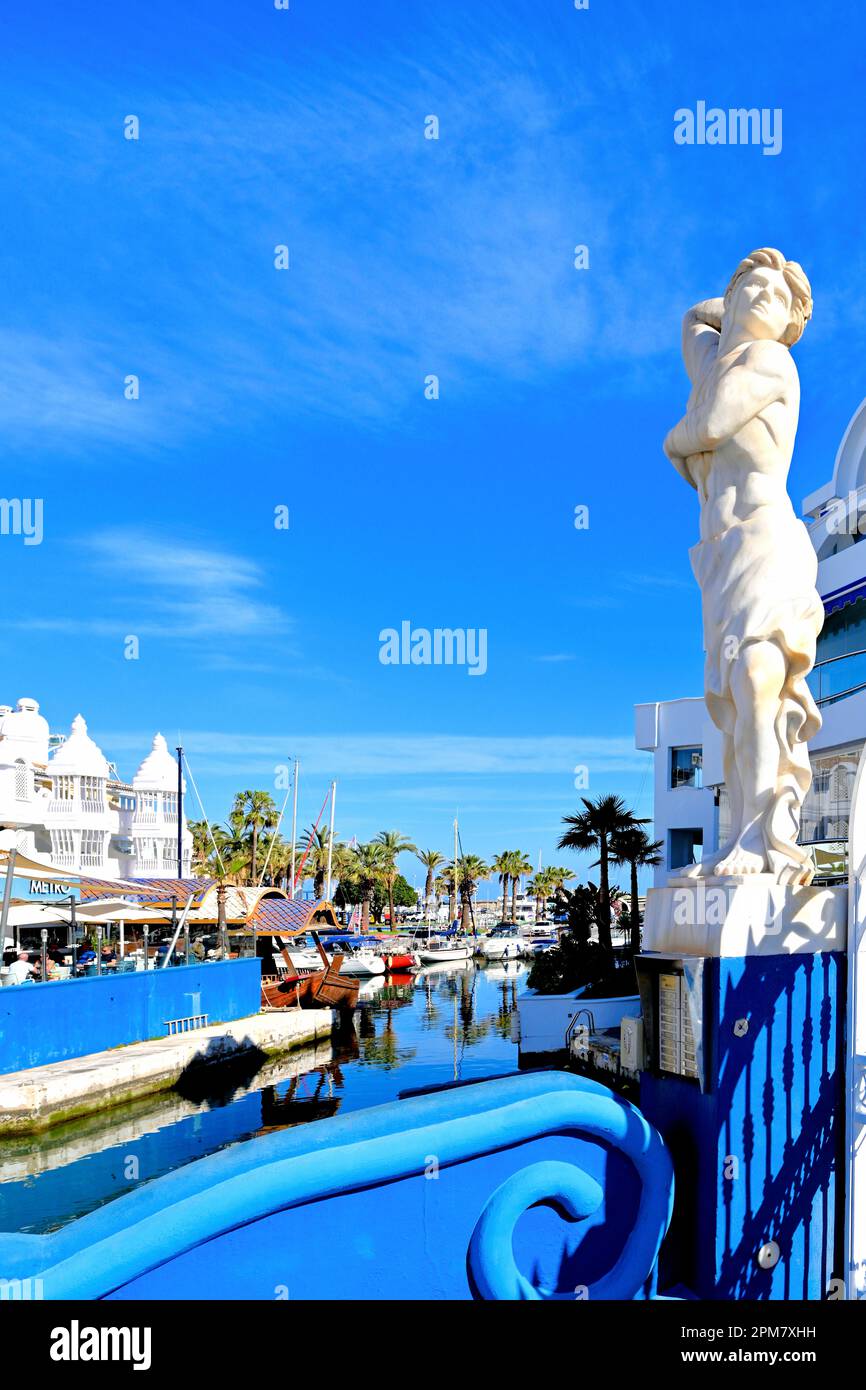 Malaga Benalmadena Marina mostra edifici bianchi con yacht e bar e una statua maschile all'ingresso contro un cielo blu profondo Foto Stock