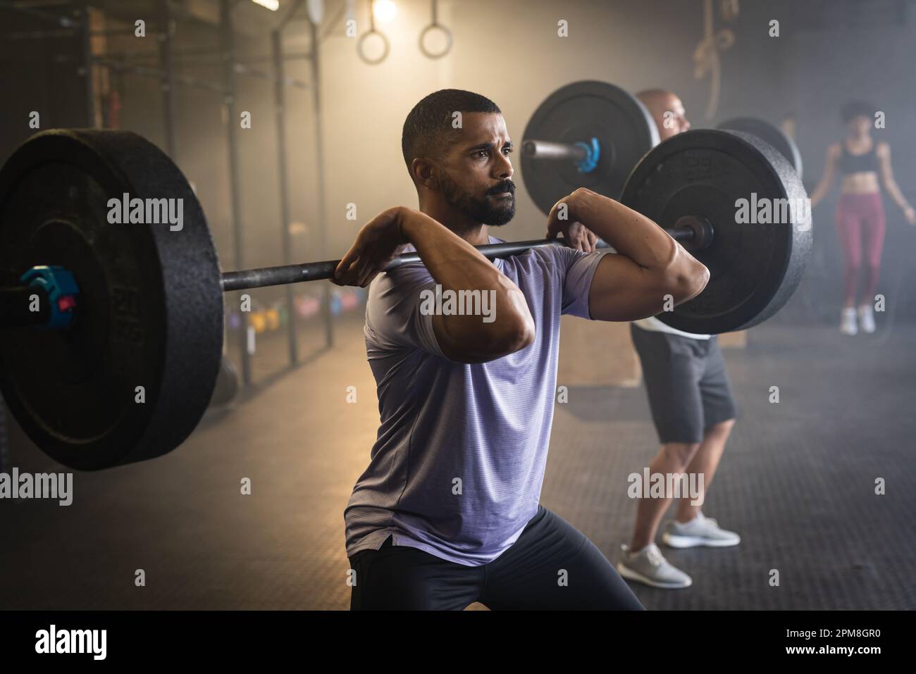 Un giovane bearded biracial dedicato che solleva il barbell mentre è in piedi in palestra Foto Stock