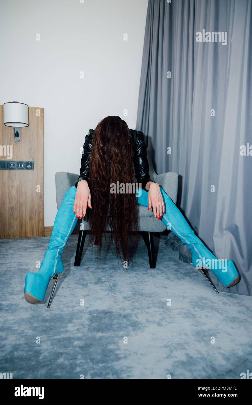 La donna che indossa stivali in pelle azzurra si siede su una sedia piegata in avanti in modo che il suo viso sia coperto dai suoi lunghi capelli scuri, che pendono in giù Foto Stock
