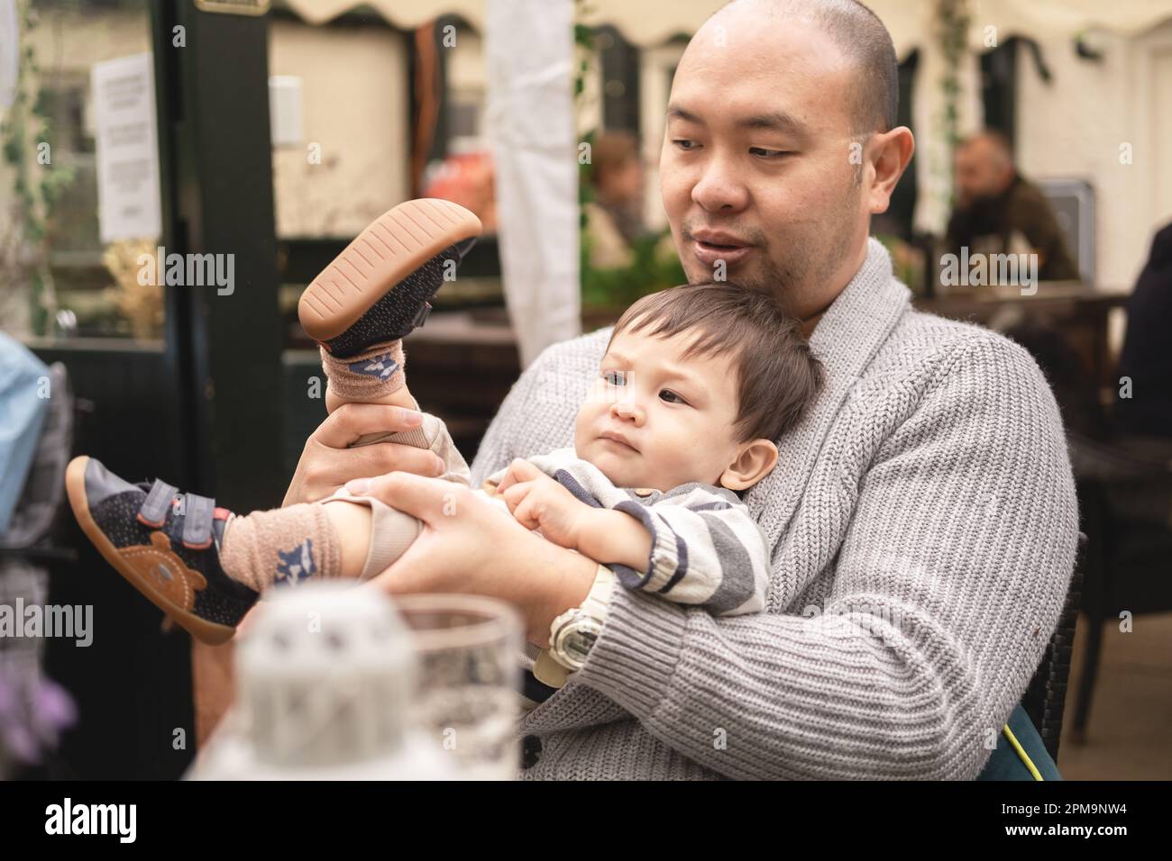 Bambino maschio flessibile seduto sulle gambe del papà, tenendo le gambe, giocando con il padre, espressione seria, in birreria all'aperto al pub in Scozia. Happ Foto Stock