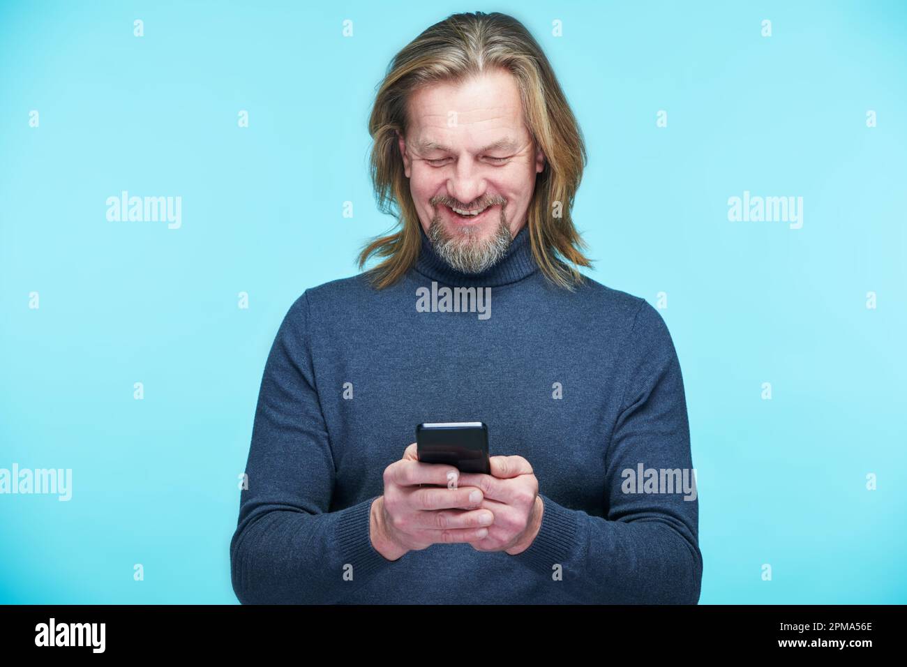 Uomo maturo che legge il messaggio sul suo smartphone e sorridente in piedi su sfondo blu Foto Stock