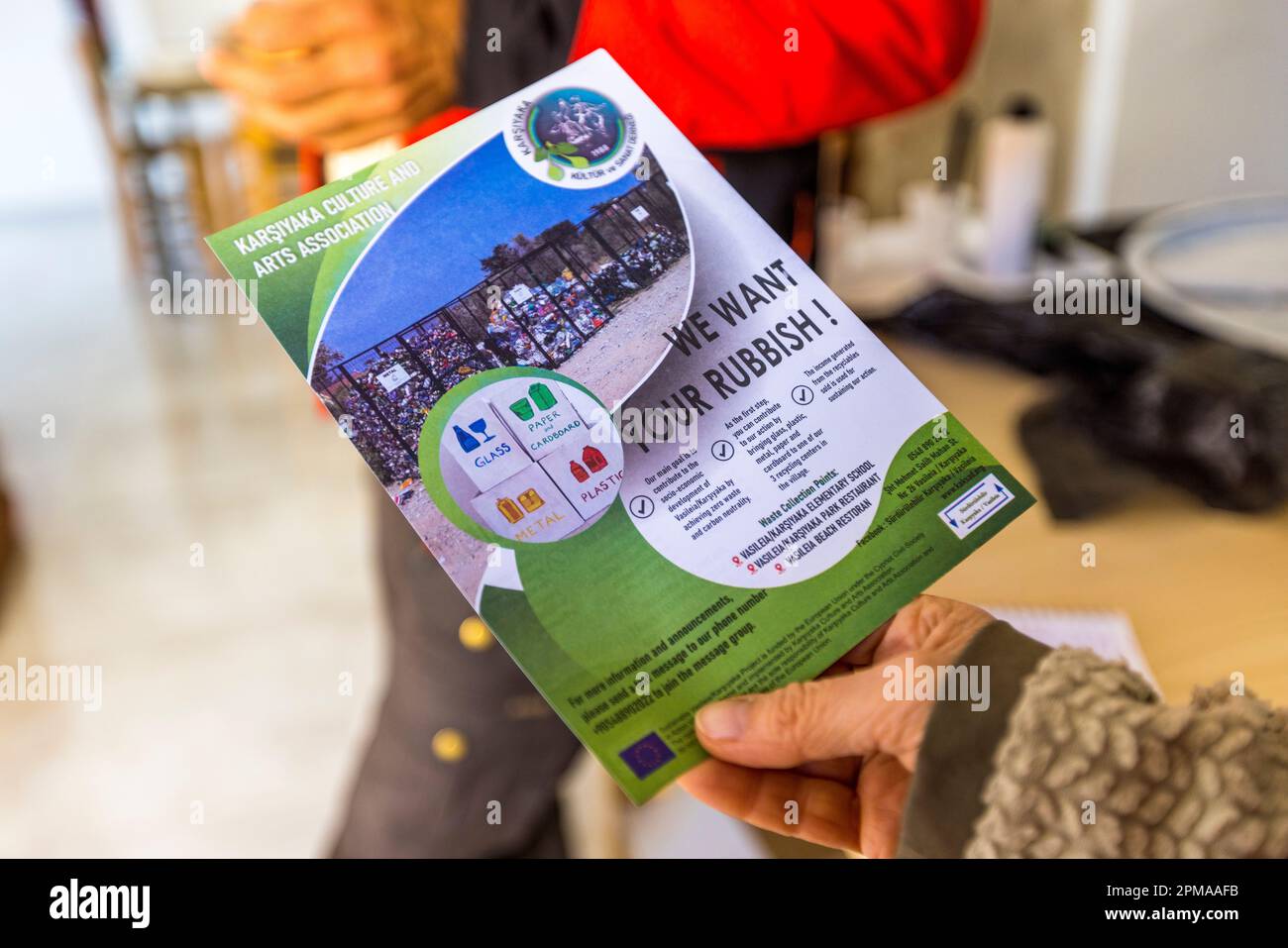 Una mano regge il volantino sul riciclo “Vogliamo i tuoi rifiuti!” dell'Associazione Culturale e Artistica di Karşıyaka a Karşıyaka (Vasilia), Repubblica Turca di Cipro del Nord (RTNC). In background, un uomo in giacca rossa spiega il progetto di riciclo. Il “Progetto Sostenibile Vasileia / Karşıyaka” include un Programma di Riciclo con l'obiettivo di “Rifiuti Zero entro il 2030”. Per raggiungere questo obiettivo, verranno organizzate serie di attività formative per i locali e gli studenti delle scuole primarie dei villaggi per insegnare loro perché la sostenibilità è importante a lungo termine. Foto Stock