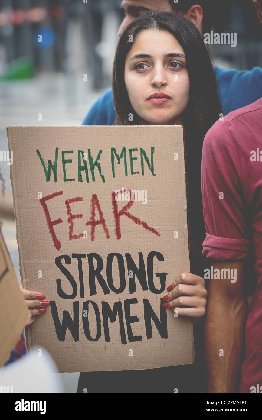 Una giovane donna mediorientale che ha un segno che dice "uomini deboli temono donne forti" durante una protesta, in piedi fuori dalla folla di altri giovani peopl Foto Stock