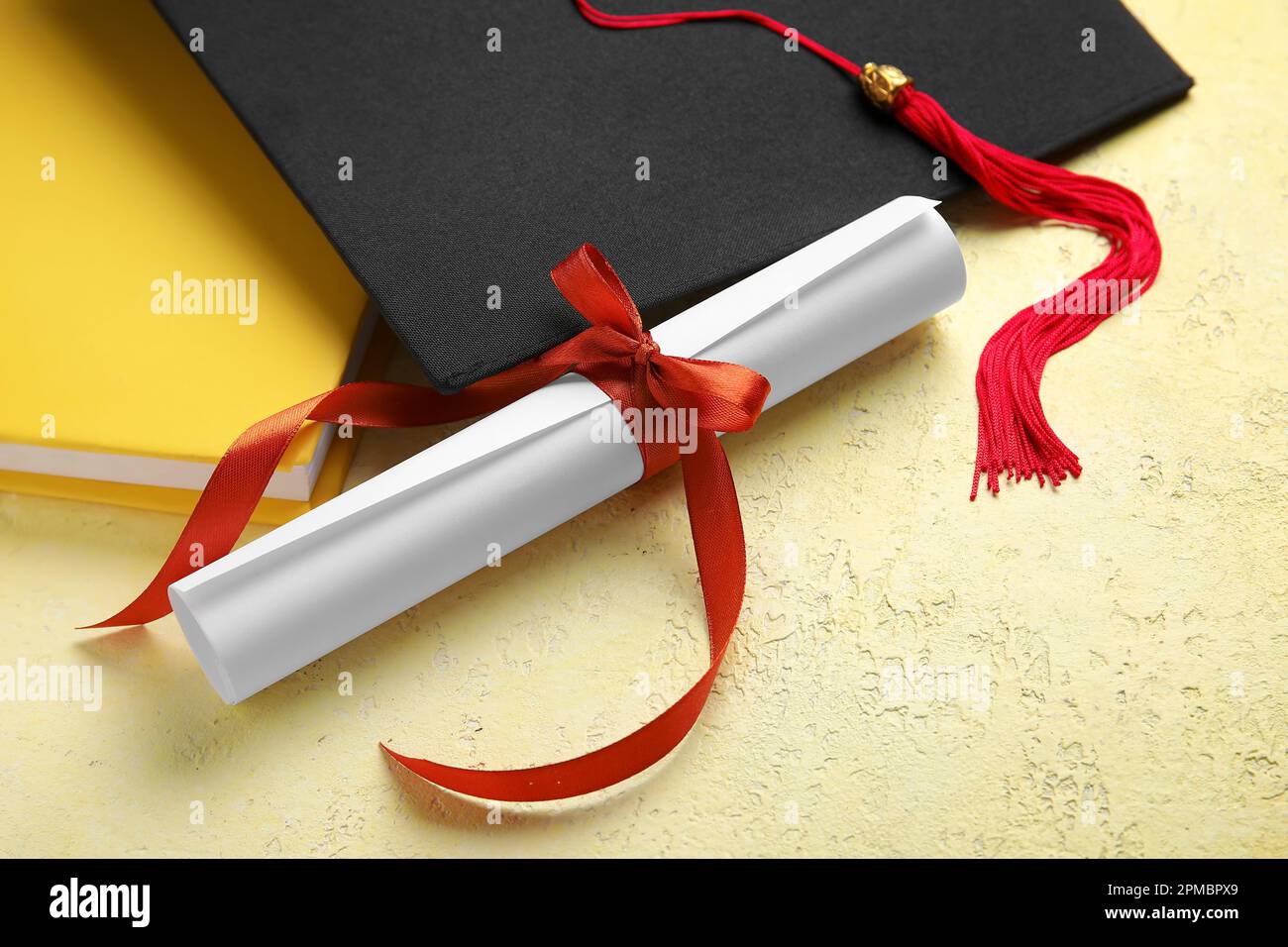 Diploma con nastro rosso, cappello di laurea e libro su tavolo giallo Foto Stock