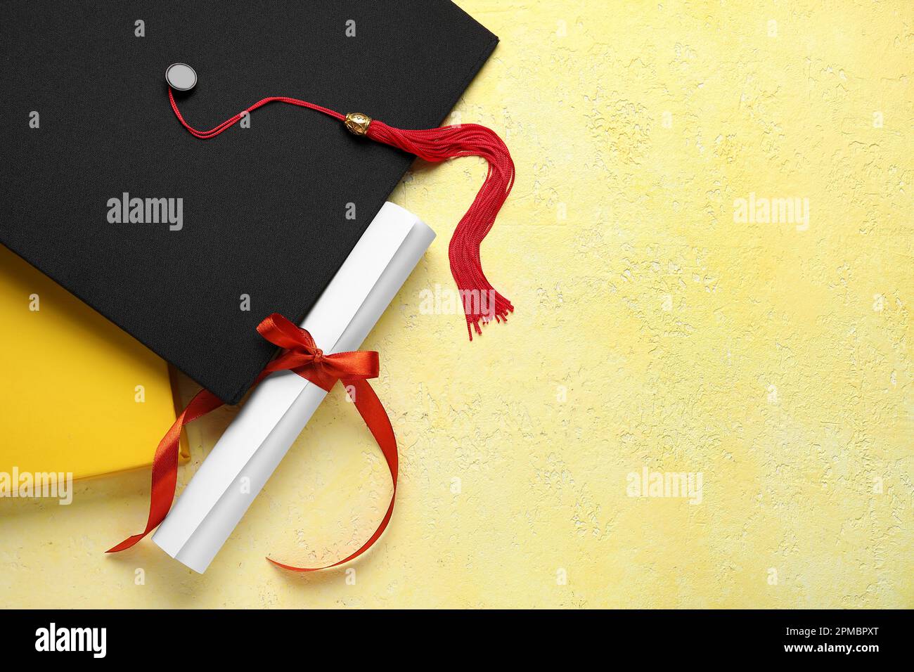Diploma con nastro rosso, cappello di laurea e libro su tavolo giallo Foto Stock
