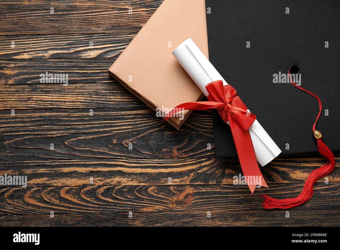 Diploma con nastro rosso, cappello di laurea e libro su tavolo di legno Foto Stock